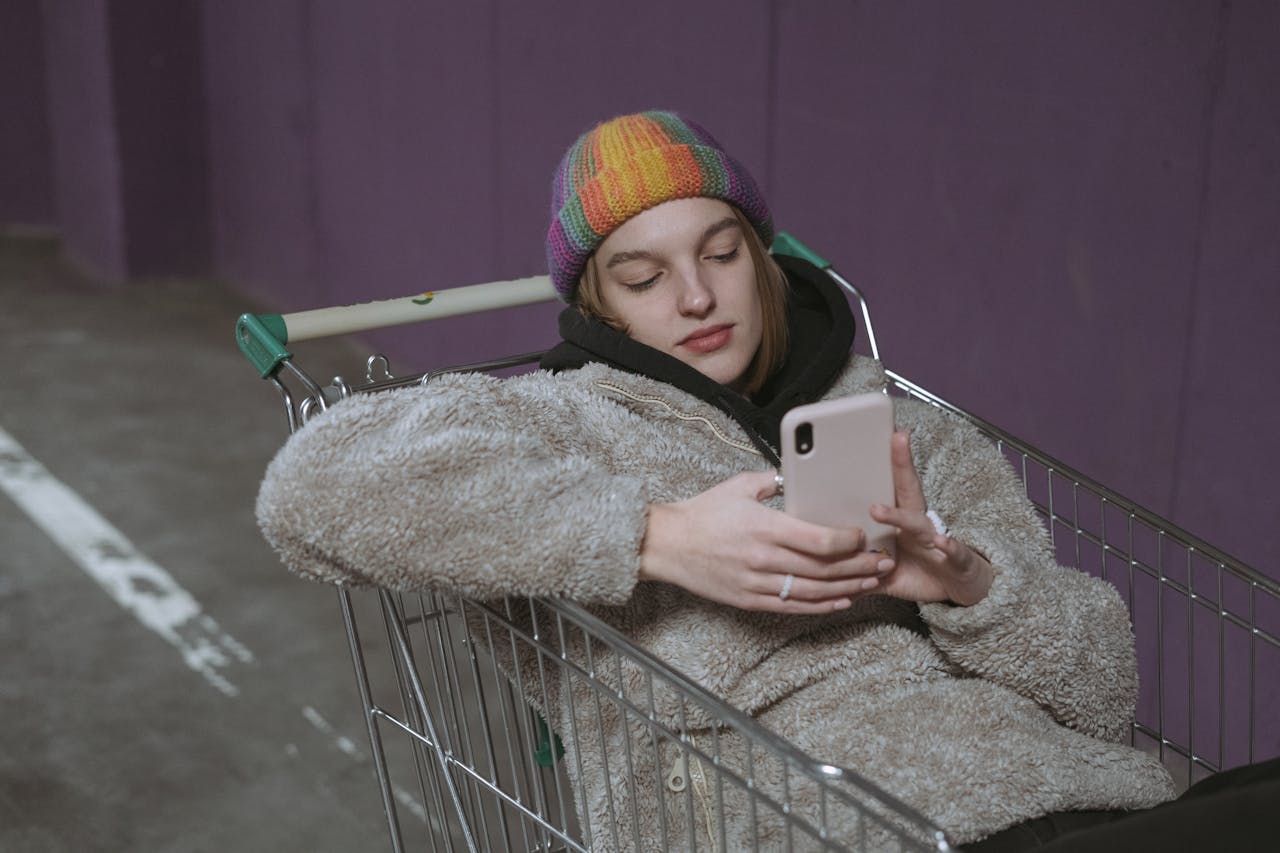 Casual scene of a woman in a beanie browsing her smartphone while sitting in a shopping cart.